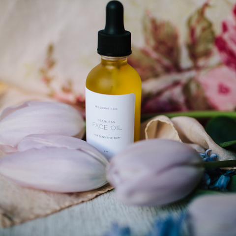 A golden oil inside of a frosted glass bottle with a black dropper top is surrounded by a vintage peach scarf with pink roses and light purple tulips and small blue flowers sit on the table