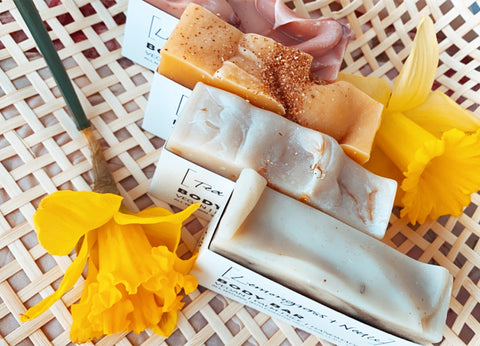 Four all-natural vegan soaps are lined up in a basket, surrounded by deep yellow daffodils. There is a pink soap, orange soap, a cream soap and a light green soap